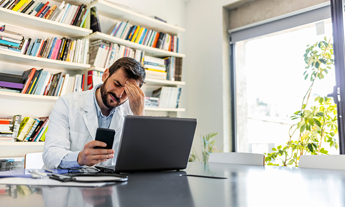Image d’un médecin assis à son bureau devant un ordinateur portable et un cahier, qui regarde son appareil mobile d’un air découragé.