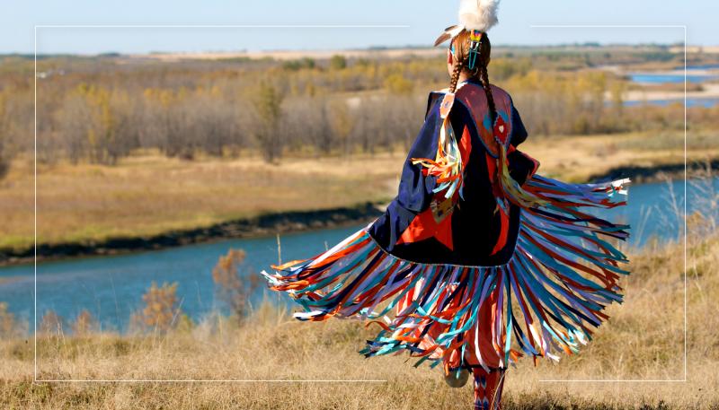 Girl in Indigenous regalia looking at a river behind her